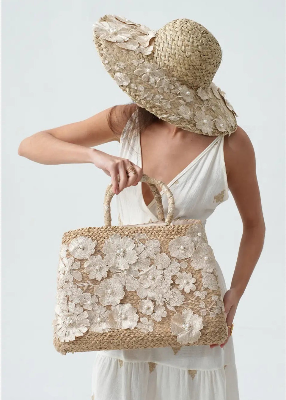 Woman holding a floral embroidered straw bag and wearing a matching hat against a light background